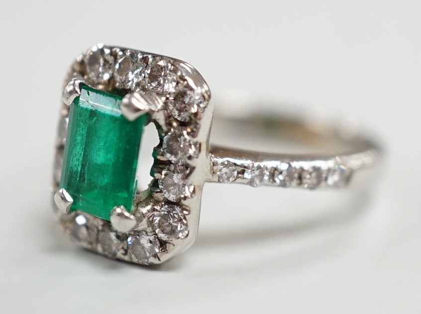 A modern 750 white metal, emerald and diamond set octagonal cluster ring, with diamond set shoulders, size J, gross weight 4.9 grams.